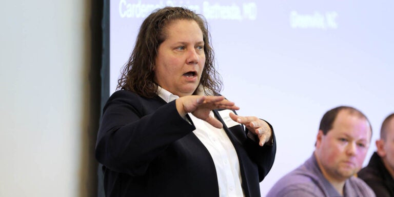 A woman in a jacket gestures with her hands as she speaks. Two man at right are seated in the background.