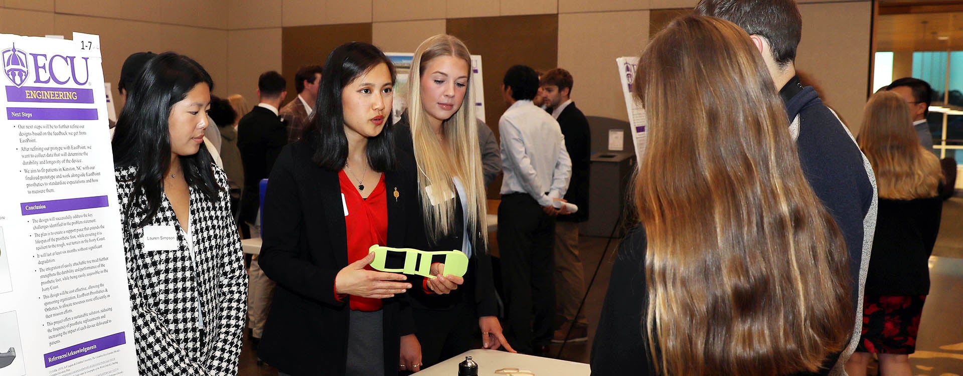 Three young women stand behind a table, with the one in the middle holding a prosthetic tread attachment, as they talk to two other people standing at right.