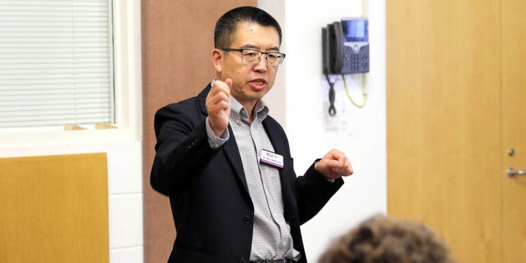 Dr. Jerry Gao, wearing a dark sport coat, gestures as he stands and speaks while a person sits at a table in front of him.