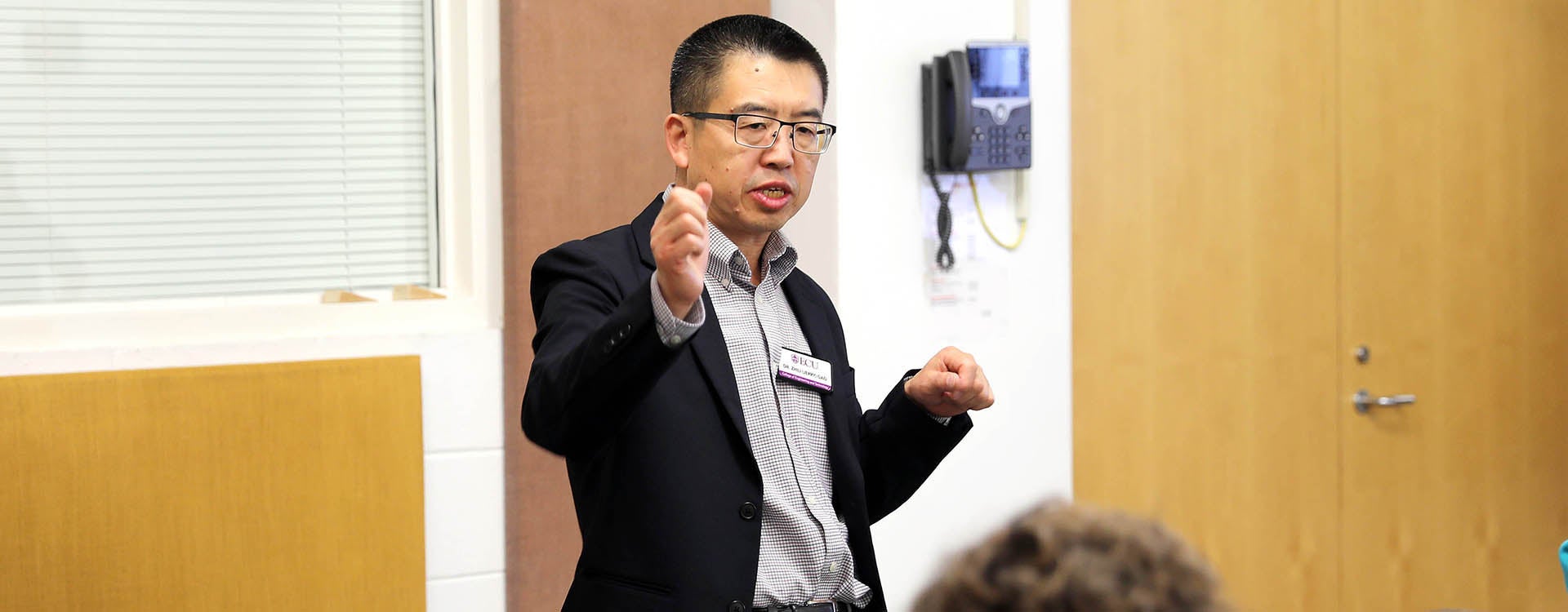 Dr. Jerry Gao, wearing a dark sport coat, gestures as he stands and speaks while a person sits at a table in front of him.