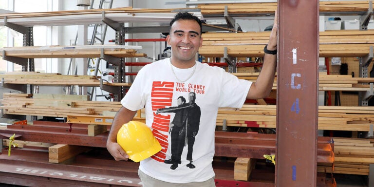 A man holding a hard hat and wearing a T-shirt stands with his hand against a steel beam with stacks of wood in the background.