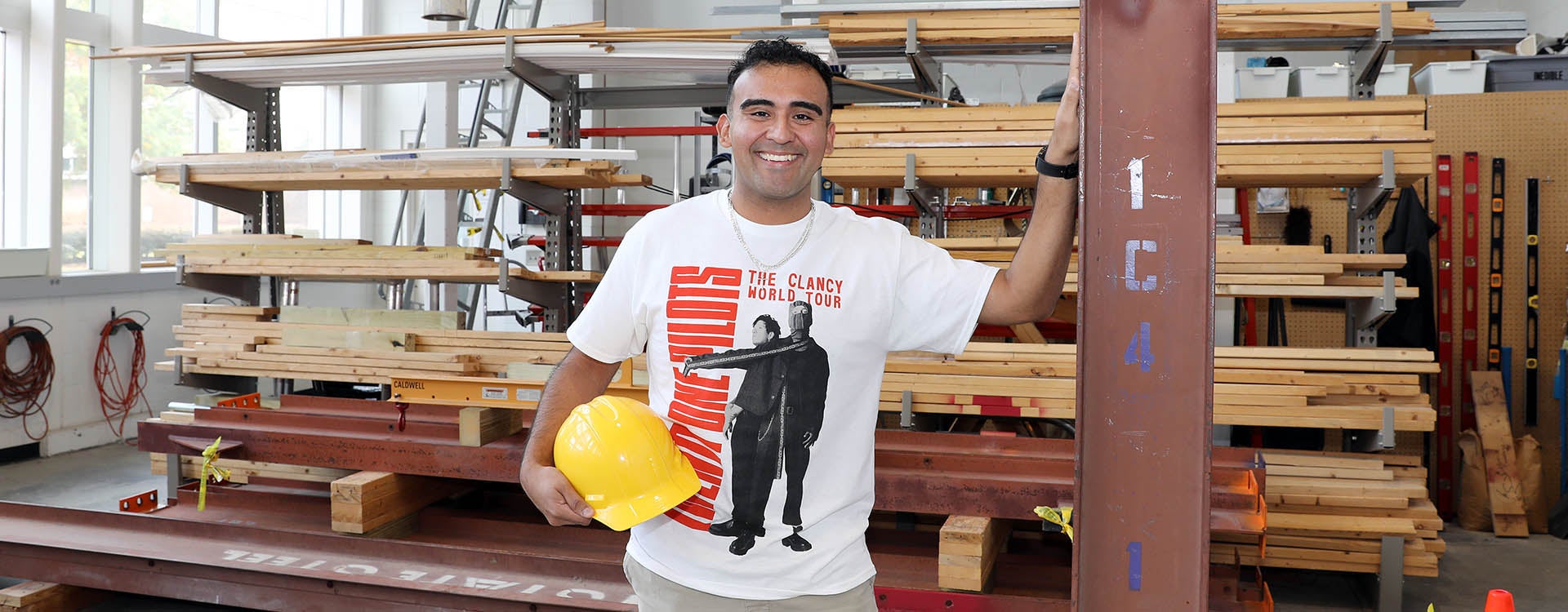 A man holding a hard hat and wearing a T-shirt stands with his hand against a steel beam with stacks of wood in the background.