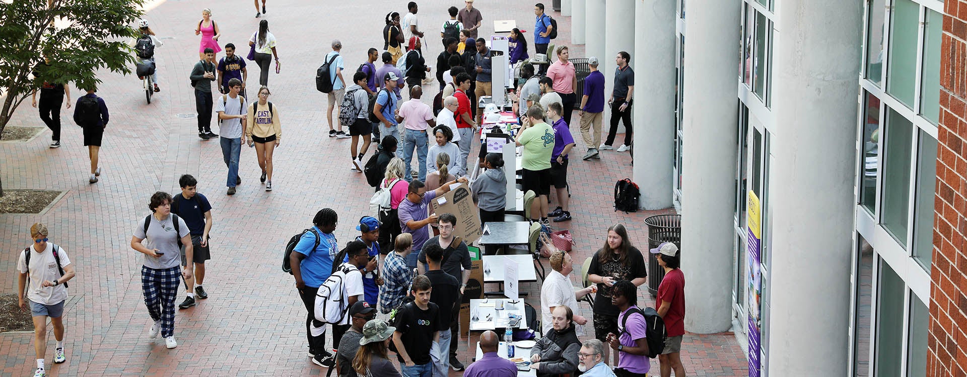 A large group of students stand and talk with others and those seated at tables as other students walk by on the left.