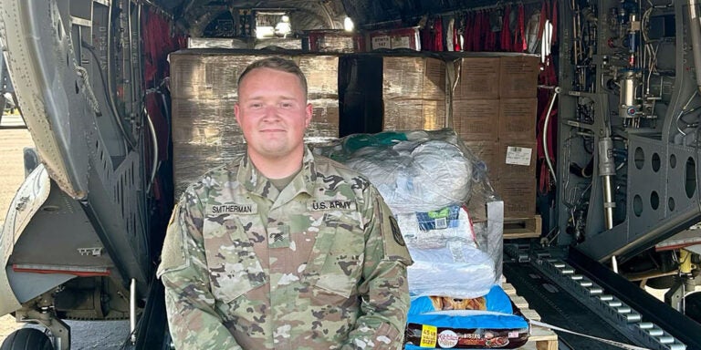 Cameron Smitherman, wearing a camouflaged uniform, stands on the ground in front of the back of an open helicopter that has pallets of boxes of storm relief supplies.