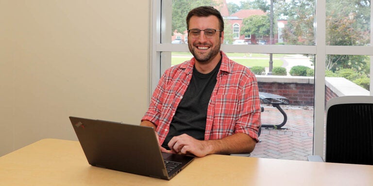 A man wearing a red shirt with a black undershirt sits at a desk in front of a laptop computer in a room with windows that show the outside.