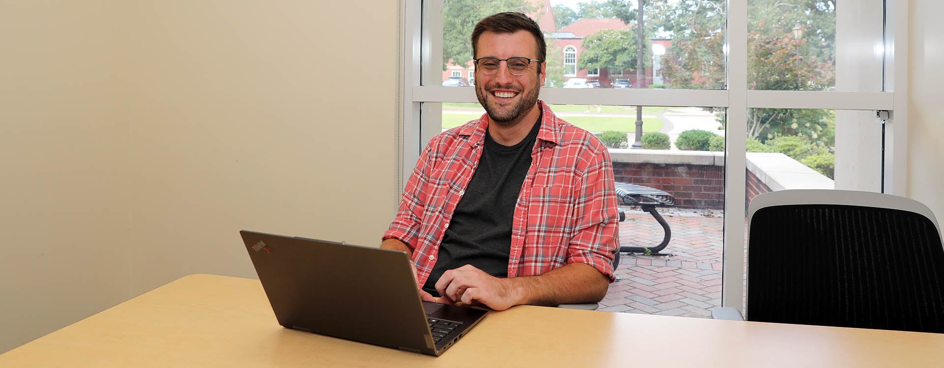 A man wearing a red shirt with a black undershirt sits at a desk in front of a laptop computer in a room with windows that show the outside.