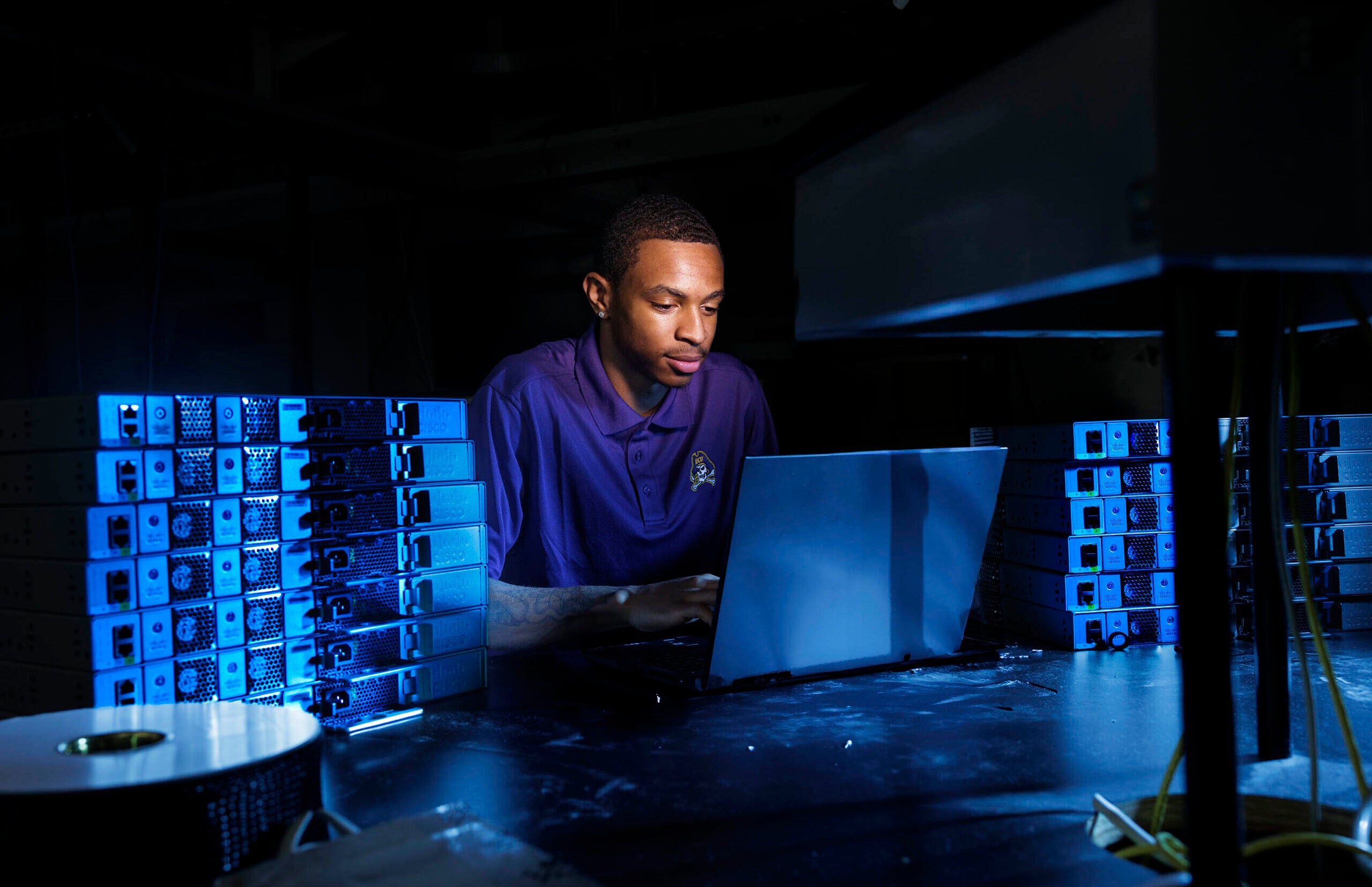 A male student wearing a purple polo with an East Carolina University logo works on a laptop on a table surrounded by stacks of computer servers.