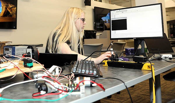 A female student with blond hair wearing a black shirt types on a computer as she sits at a table covered with various wires and computer parts.