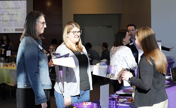 Two women at left stand in front of a display table and talk to another woman standing behind the table.