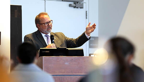 A man in a suit and tie gestures with his hand as he speaks from a podium as others sit and listen.