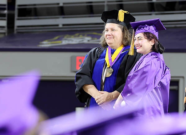 A woman at left in a black graduation cap and gown stands next to a younger woman in a purple graduation cap and gown.