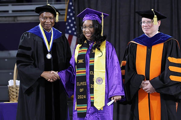 A young woman in a graduation cap and gown stands between two men in graduation robes as they pose for a picture on a stage.