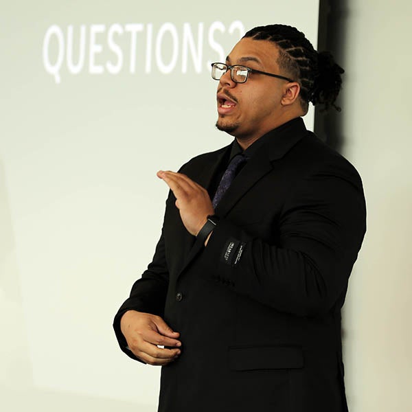 A man in a dark suit gestures with his hands as he speaks in front of a large projection screen with the word questions on it.