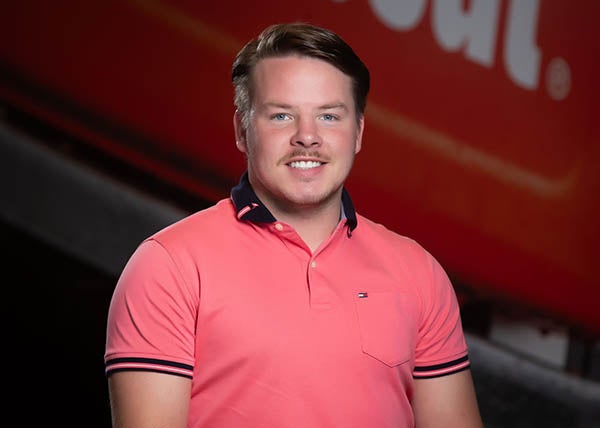 A man in a collard shirt smiles and poses for a picture against a red backdrop.