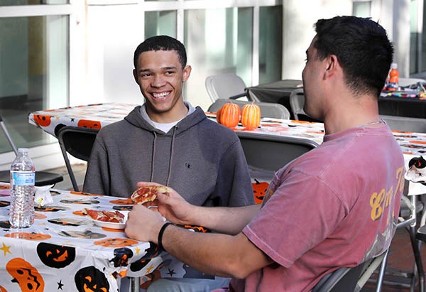 A young man in a gray hooded shirt sitting at a table smiles while talking to another young man wearing a T-shirt sitting at the right.