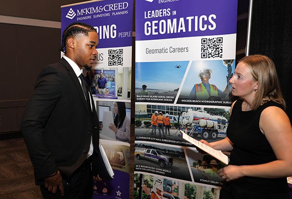 A man at left in a suit and tie talks to a woman wearing a black shirt in front of a banner for McKim & Creed.