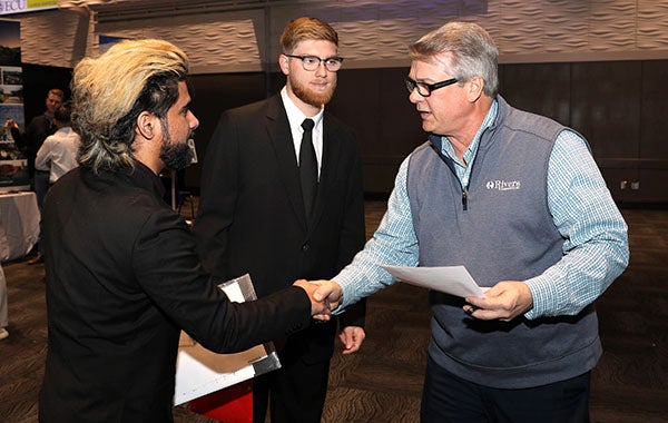 A man in a sport jacket at left shakes hands with another men in blue shirt and sweater vest at right while another man in a tie and jacket stands in the background in the center.