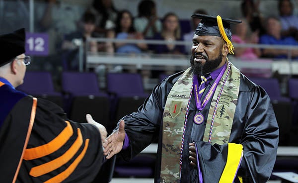 A man at right in a black graduation gown and cap reaches out to shake hands with another man in a graduation robe on a stage.