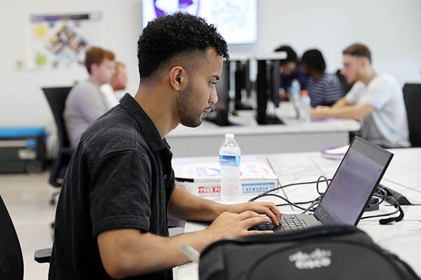 A college student in a black shirt types on a laptop computer while sitting at a long desk. Other students in the background type on computers as well.