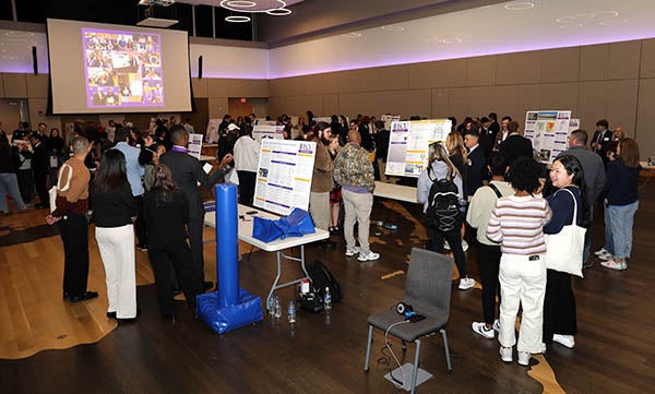 A large group of people stand I a big room full of tables, poster displays and exhibits.