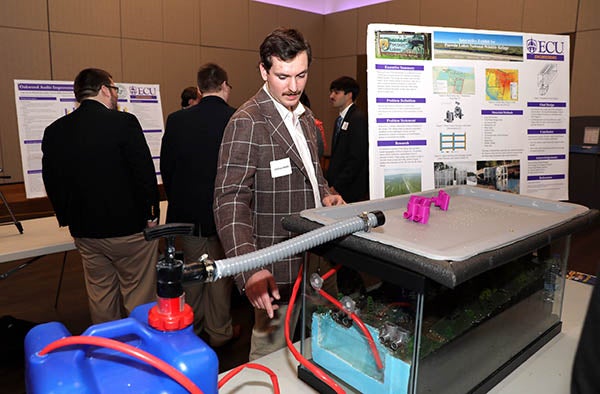 A young man in a brown jacket stands at a table behind a small blue tank to the left with a tube that leads to an aquarium tank that has a display of the ground with trees and waterways.