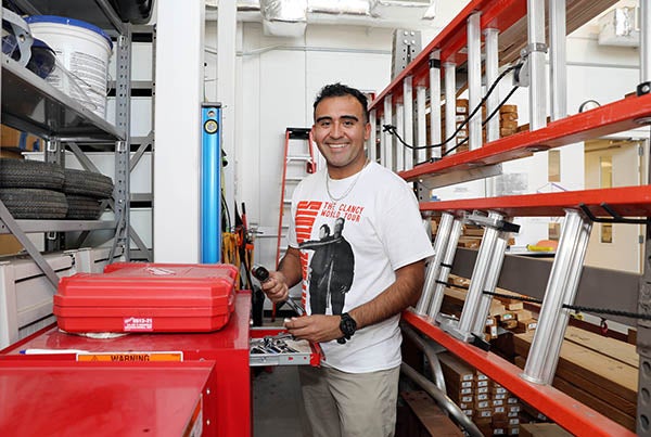 A man in a white T-shirt stands next to a tool box with a ladder to his side as he holds a wrench.