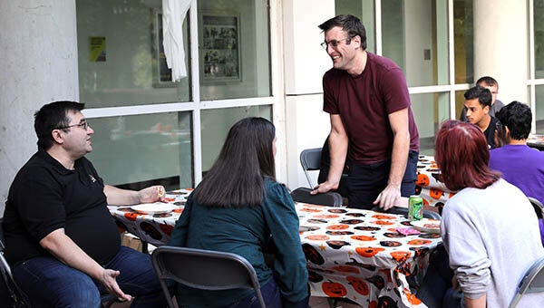 A man in a red shirt stands at a table outside and talks to three people sitting at the table.