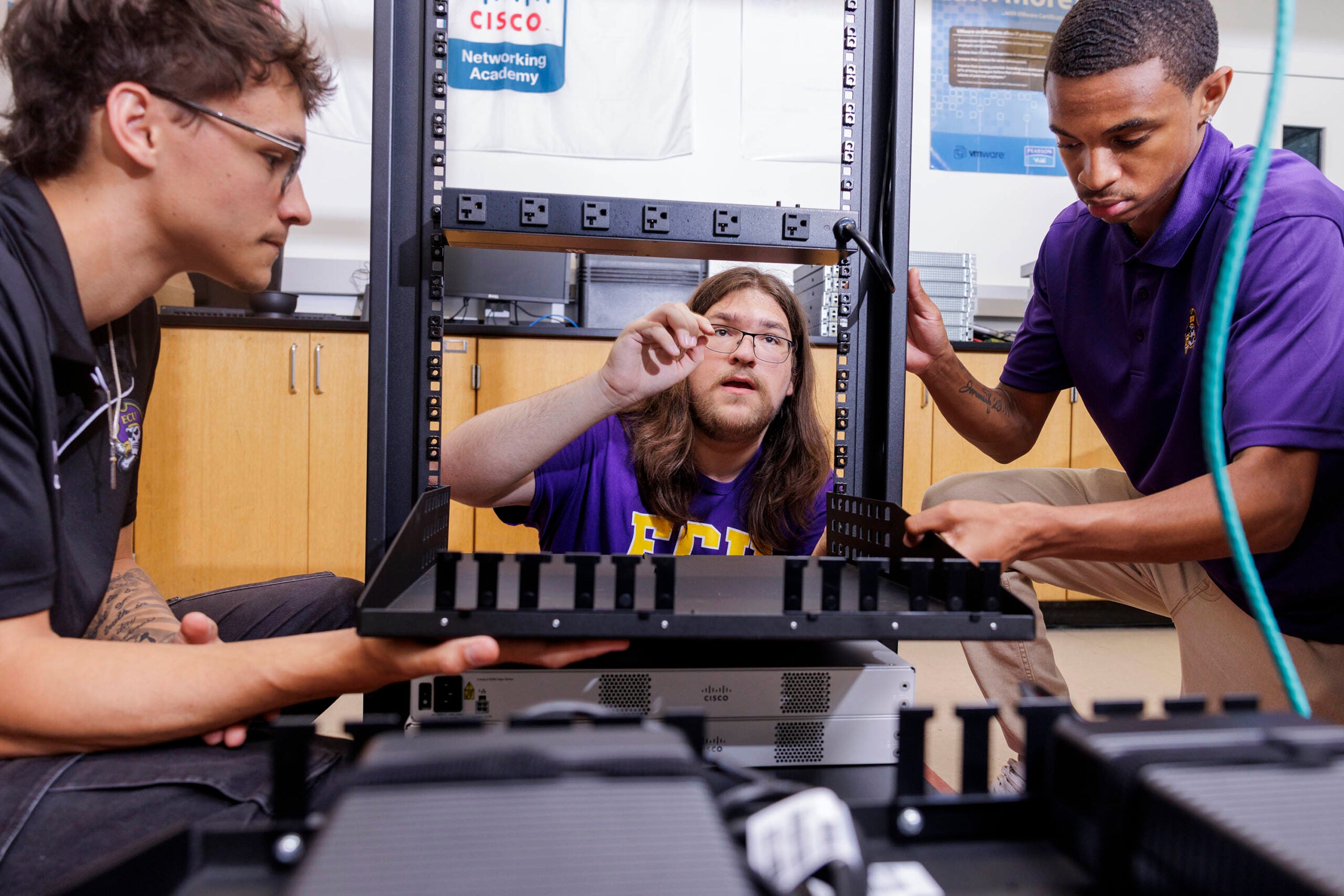 Three young men surround a vertical black, metal rack, with the men on the left and right holding a shelf while the man at center points to a location on the rack where it will be installed.