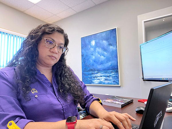 Ana Craig Harmon, wearing a purple shirt and sitting in a chair at a desk, types on a computer in an office.