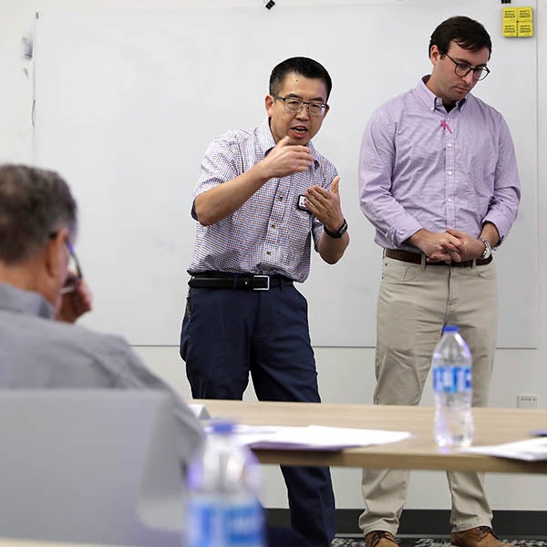 Dr. Jerry Gas stands in the front of a table where one person is sitting and next to another man who is standing and gestures with his hands as he speaks.