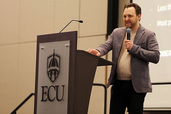 A man in a brown jacket holds a microphone as he stands next to a podium and speaks.