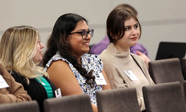 Three young women, one at left in a black sweater, one in the middle with a flowered shirt and one at right in a brown sweater, sit in chairs and listen to a presentation. The woman in the middle is smiling.